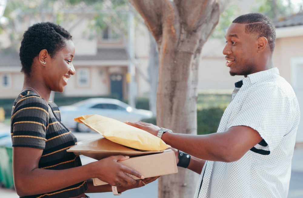 A smiling woman hands packages to a smiling man outdoors, with houses and a tree in the background on a sunny day.