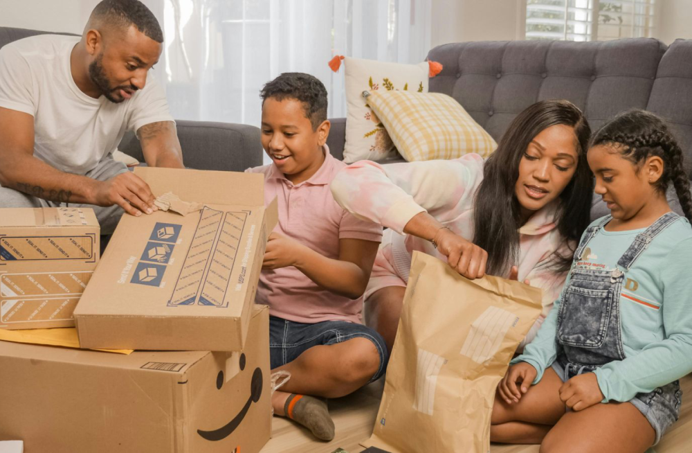 A family of four sits on the floor in a living room, happily opening packages together. Boxes and padded envelopes are spread out, and everyone appears engaged and joyful.